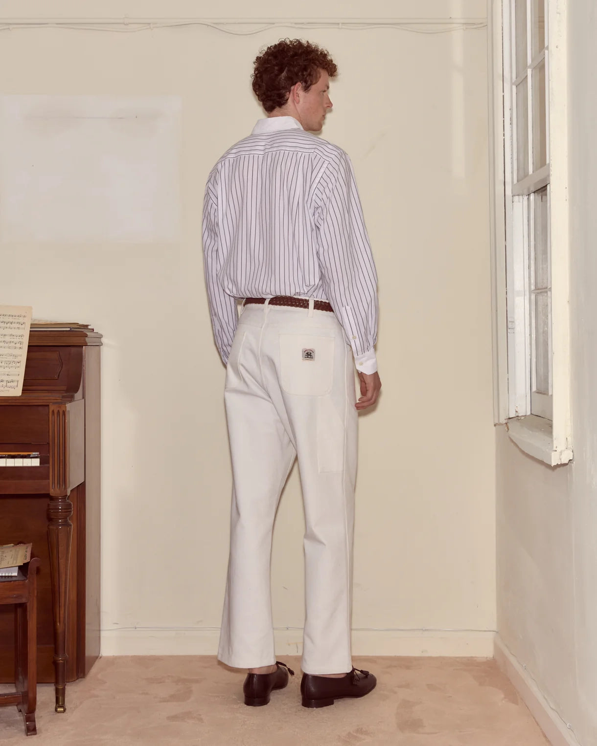Man wearing a striped shirt and white pants standing in a room with a piano.