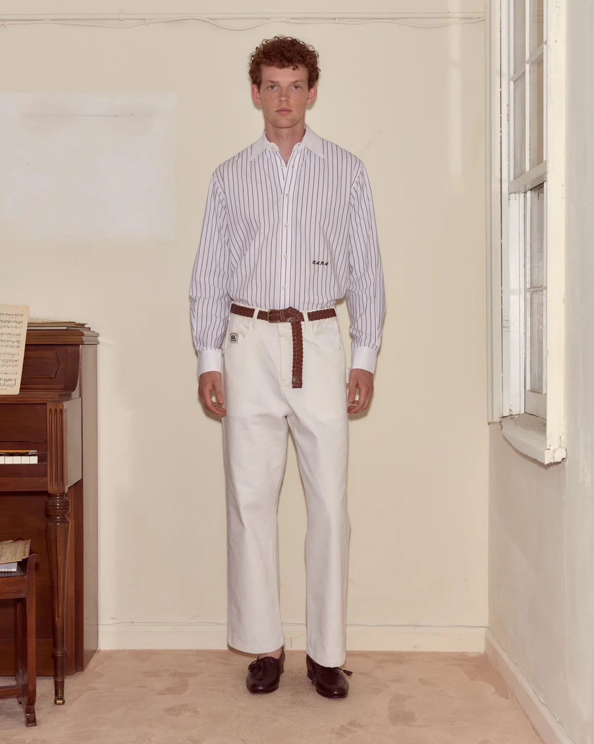 Man wearing a striped shirt and white pants standing in a room with a piano.
