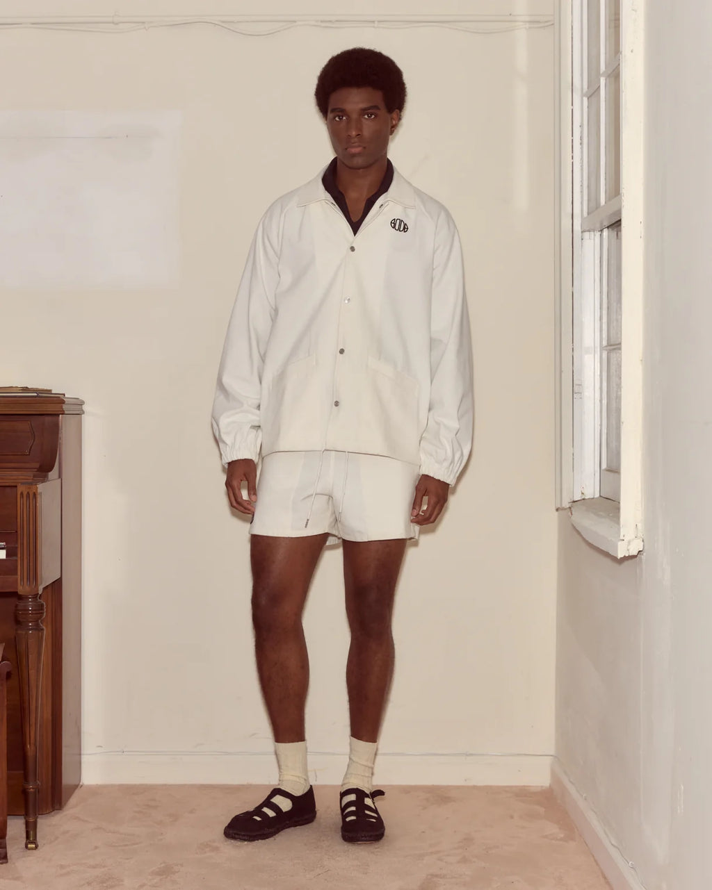 Man wearing a white shirt and shorts in a room with a window and wooden furniture.
