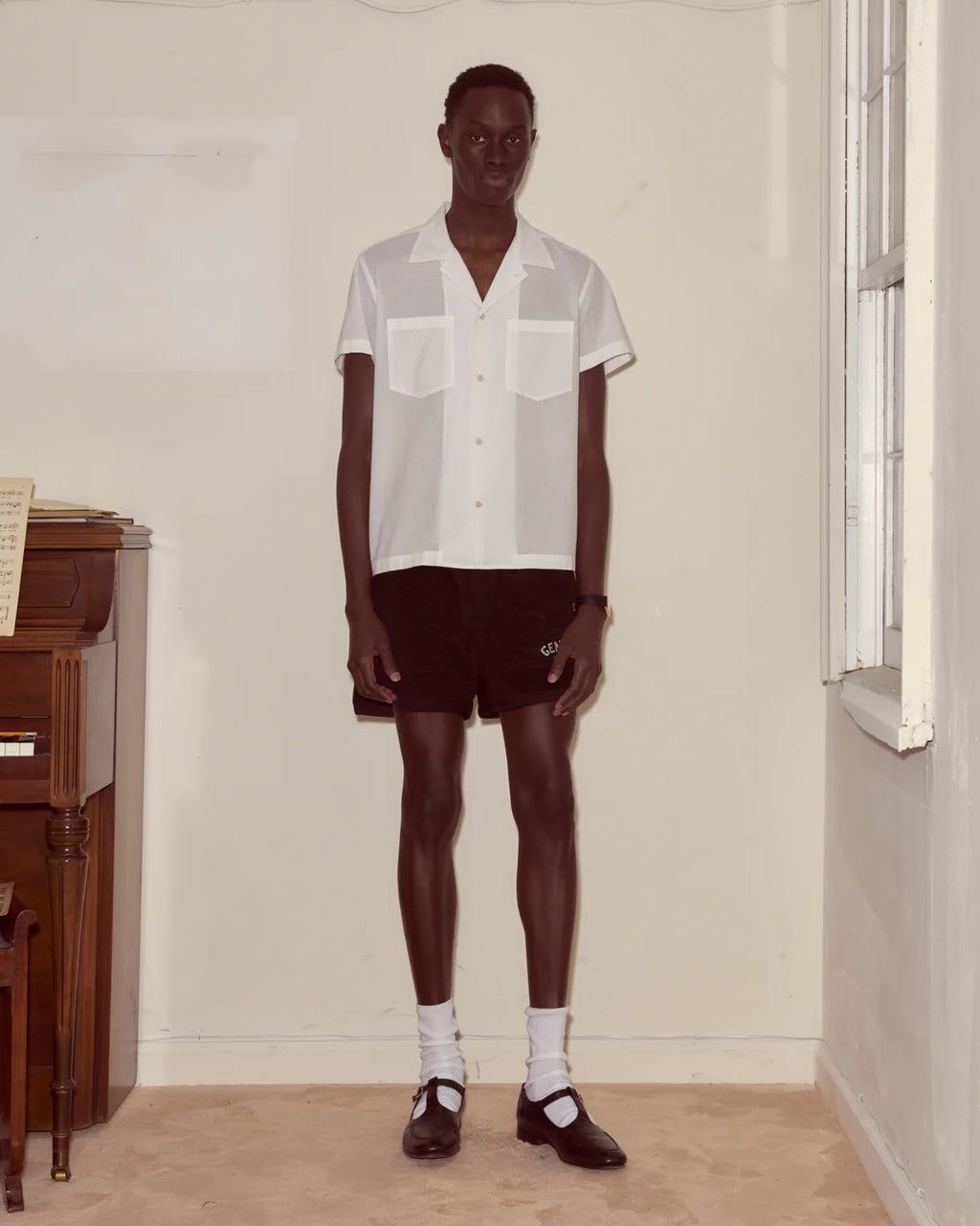 Man wearing a white shirt and dark shorts standing in a room with a window and wooden furniture.