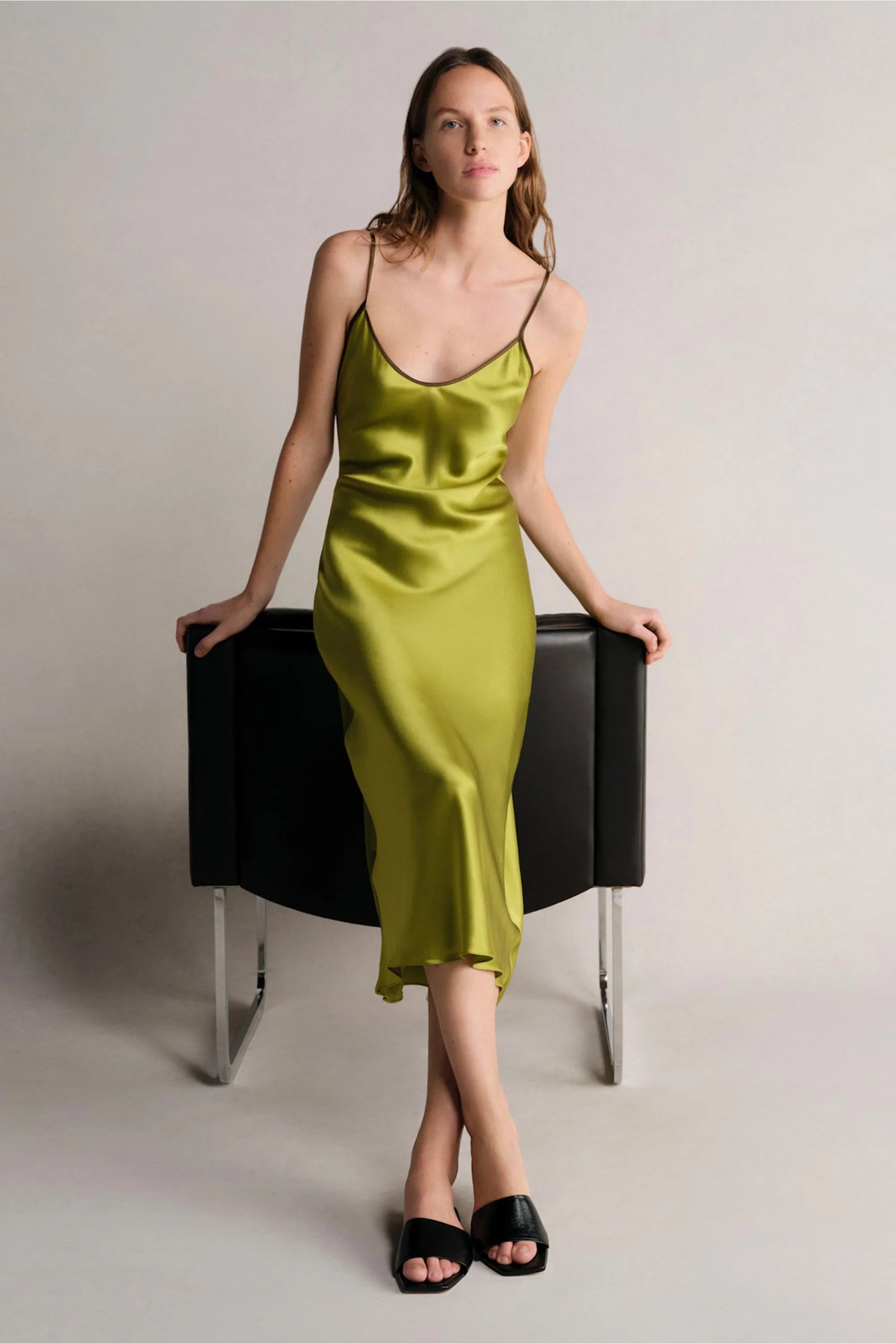 Woman wearing a green satin slip dress sitting on a black stool against a plain background