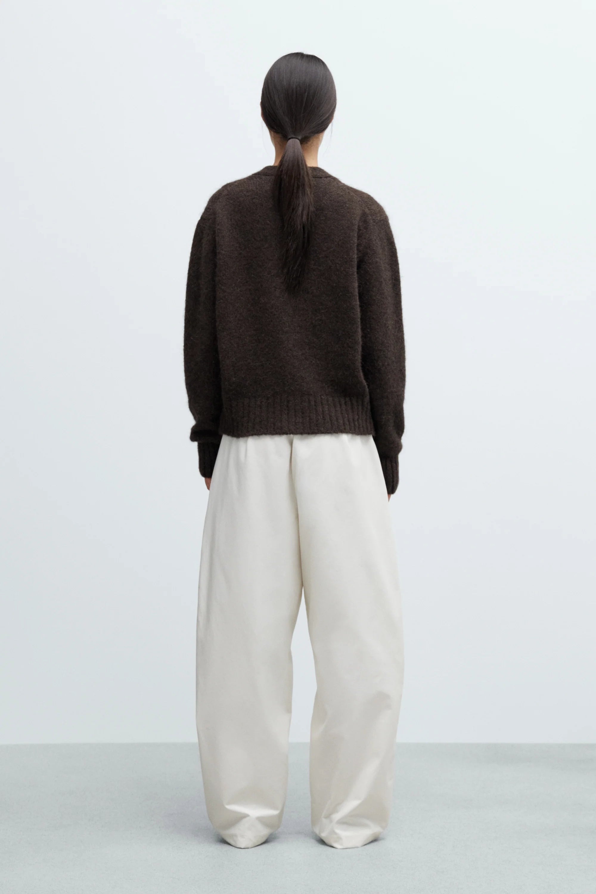 Person wearing a brown sweater and white pants on a light gray background