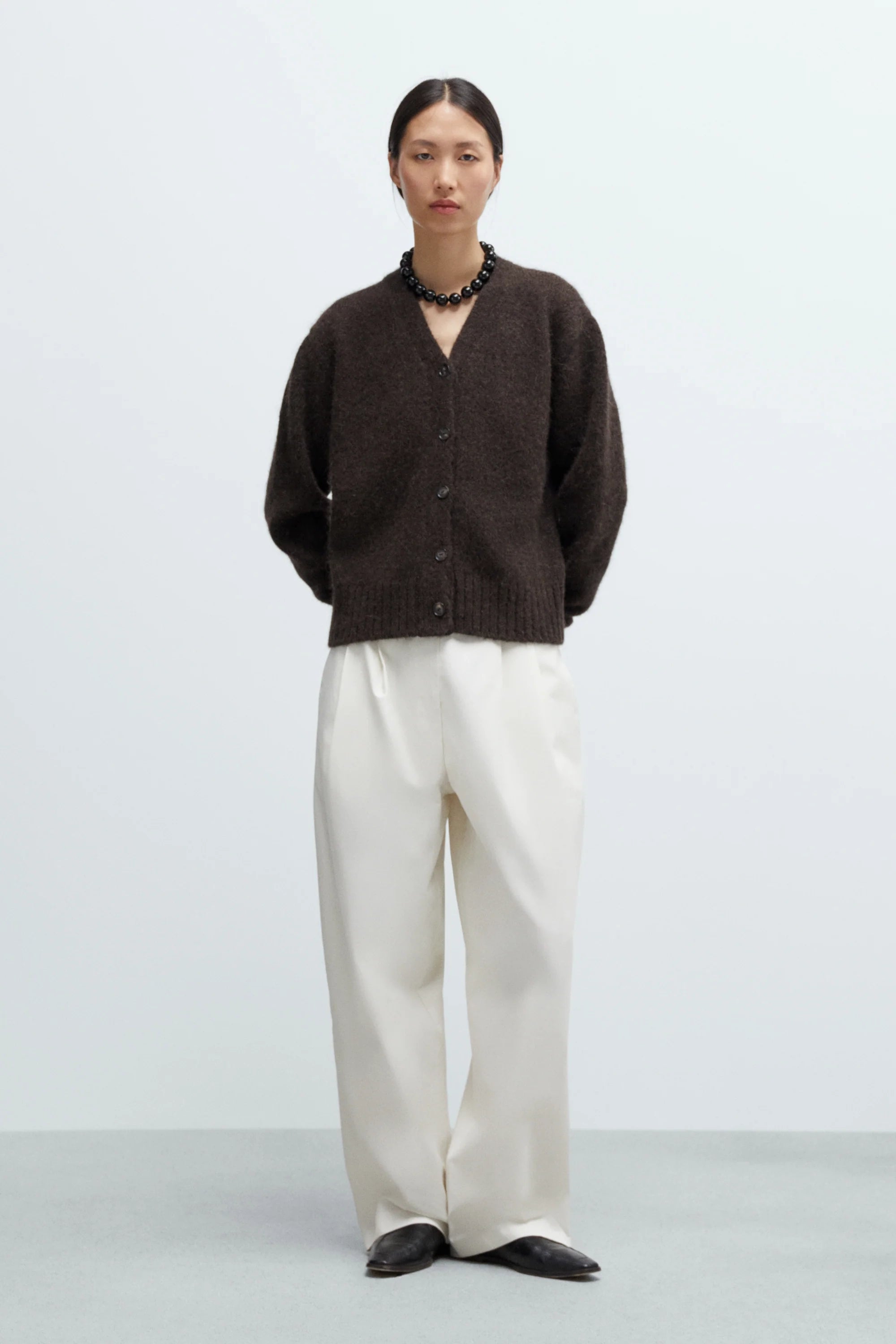 Person wearing a brown cardigan, white pants, and black shoes on a light gray background