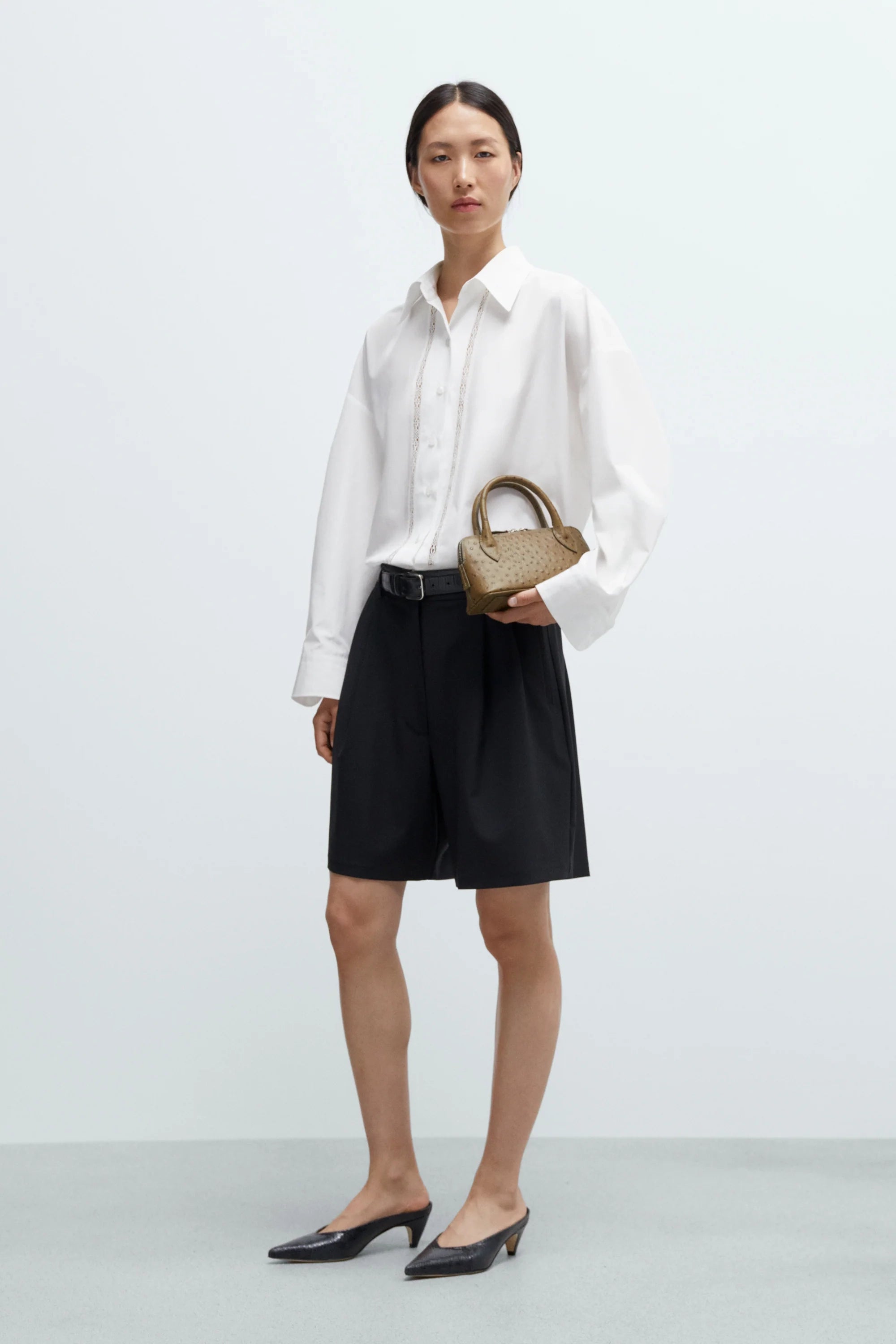Woman wearing a white shirt and black shorts holding a woven handbag on a plain background