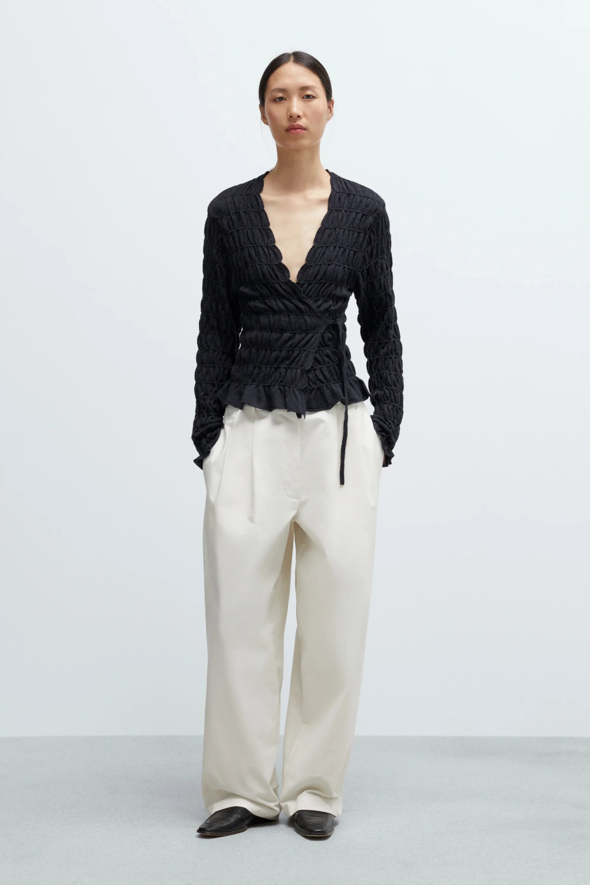 Person wearing a black textured top and white pants on a light gray background