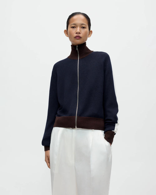 Model wearing CORDERA Merino Wool Sporty Cardigan in navy with white sleeve stripes, brown ribbed trim and a front zip closure.