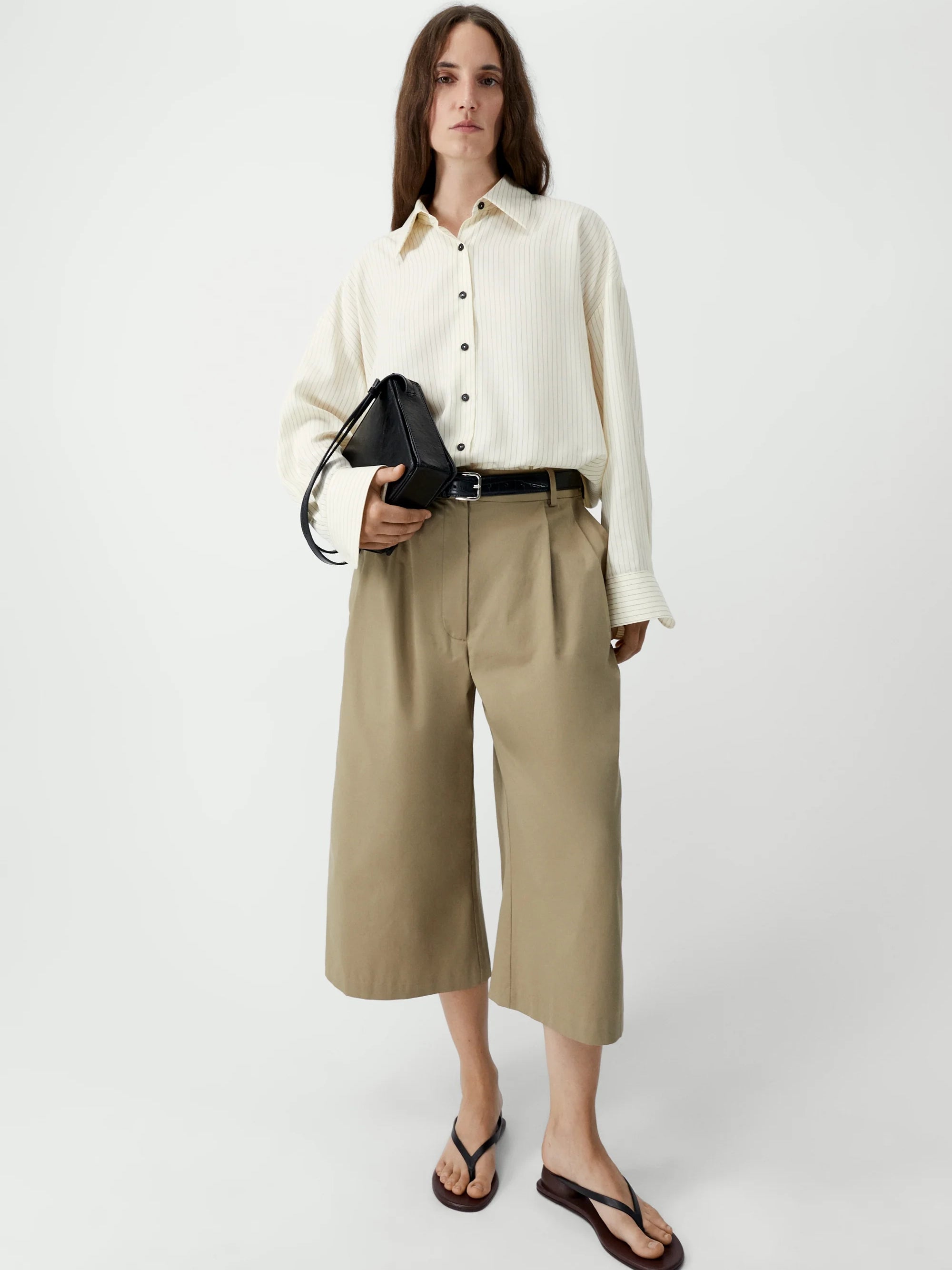 Woman wearing a white shirt and beige pants holding a black handbag on a white background