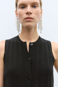 Person wearing a black sleeveless top with buttons and long earrings on a light gray background