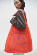 A person wearing a grey shirt and orange long earrings, holding a vivid orange shopping bag with a blue embroidered Galicia map and the brand name 'Cordera'.