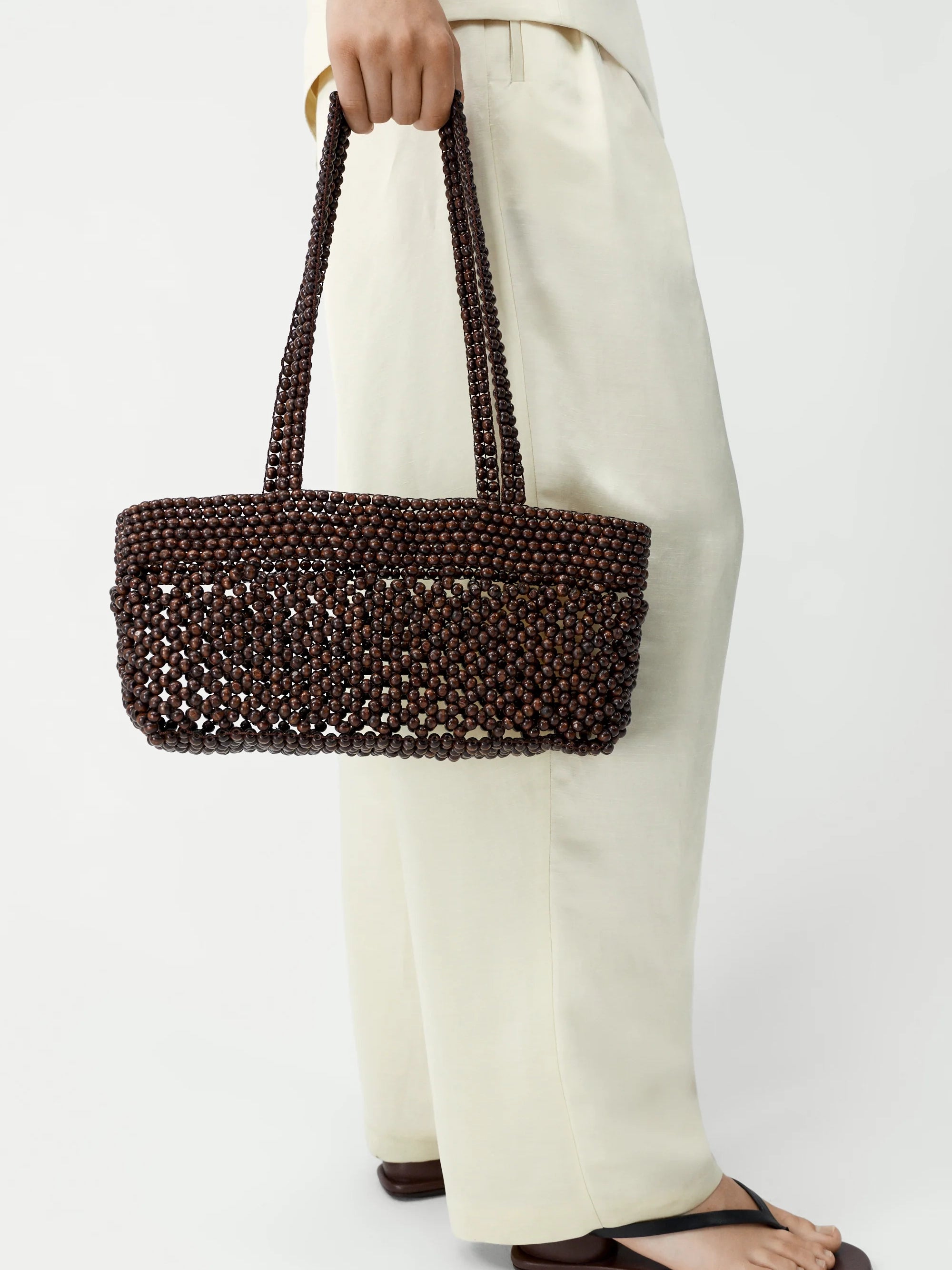 Brown beaded handbag held by a person wearing white pants on a white background