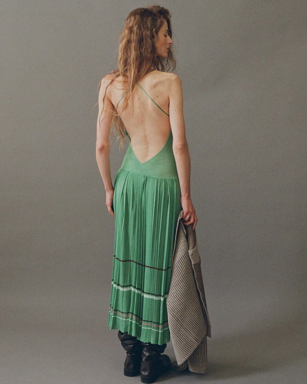 Model wearing lily pad green pleated midi dress by EMILY DAWN LONG with an open back, fine shoulder straps and striped pleated skirt.