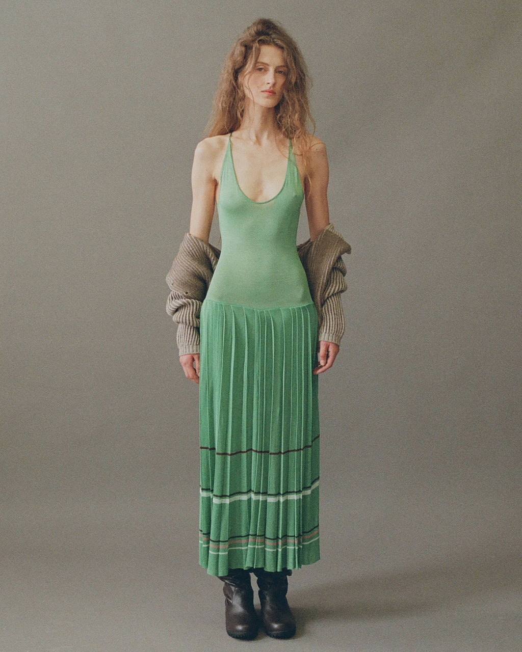 Model wearing lily pad green pleated midi dress by EMILY DAWN LONG with an open back, fine shoulder straps and striped pleated skirt.
