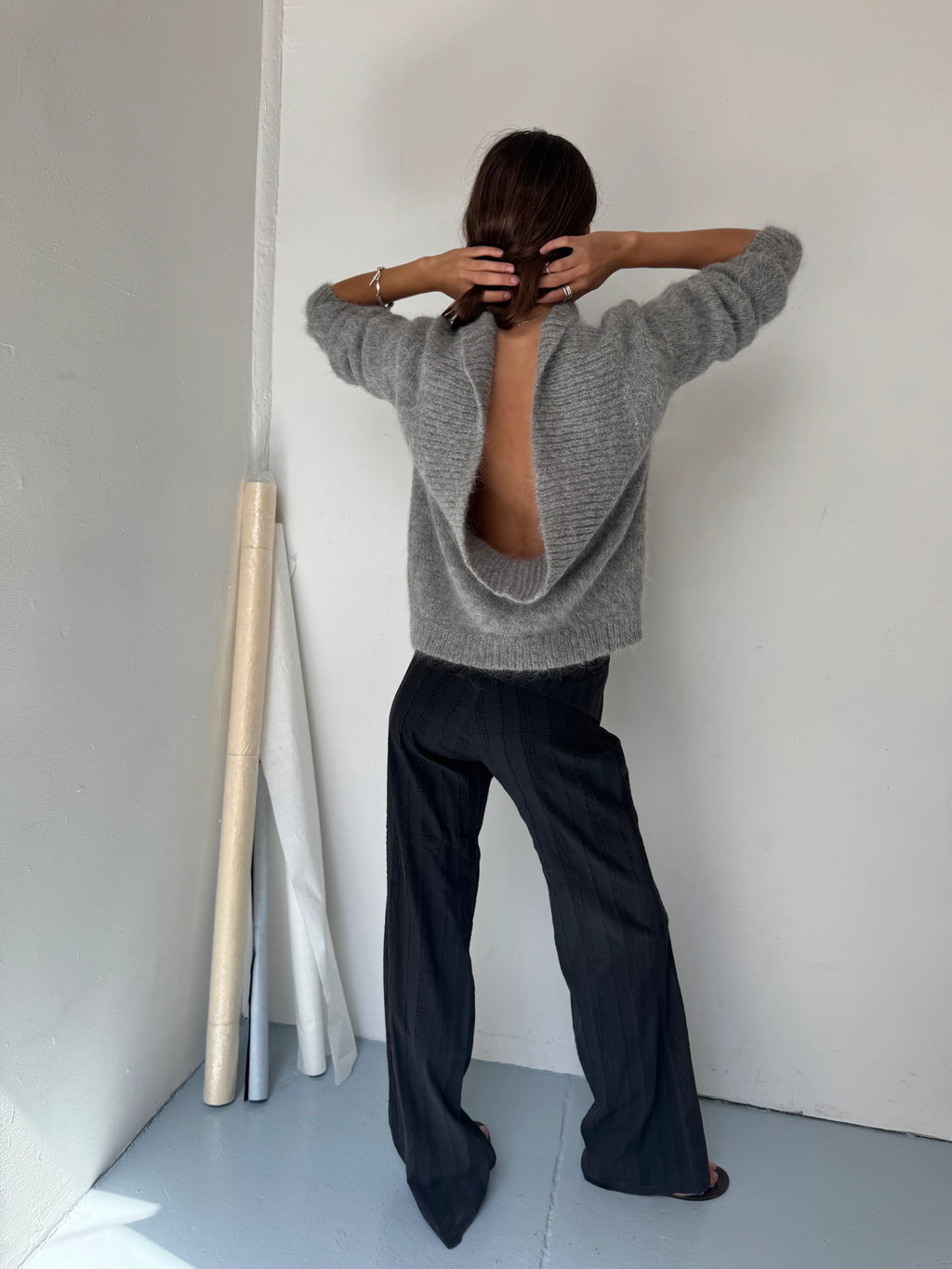 Person wearing a gray sweater and black pants standing in a minimalistic room.