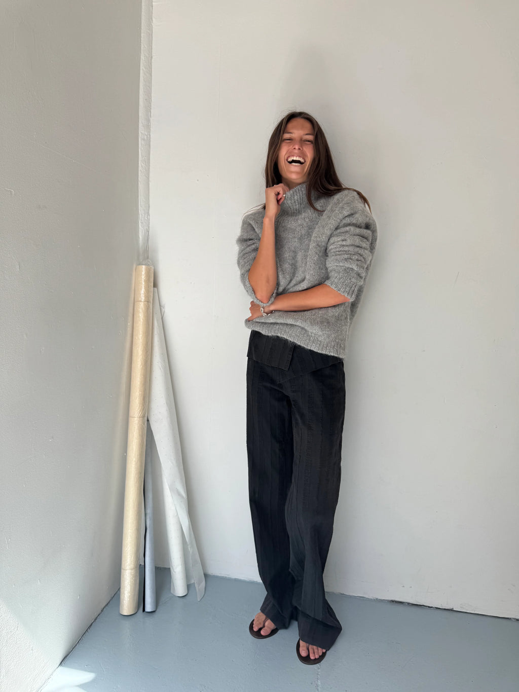 Person wearing a gray sweater and black pants standing against a white wall.