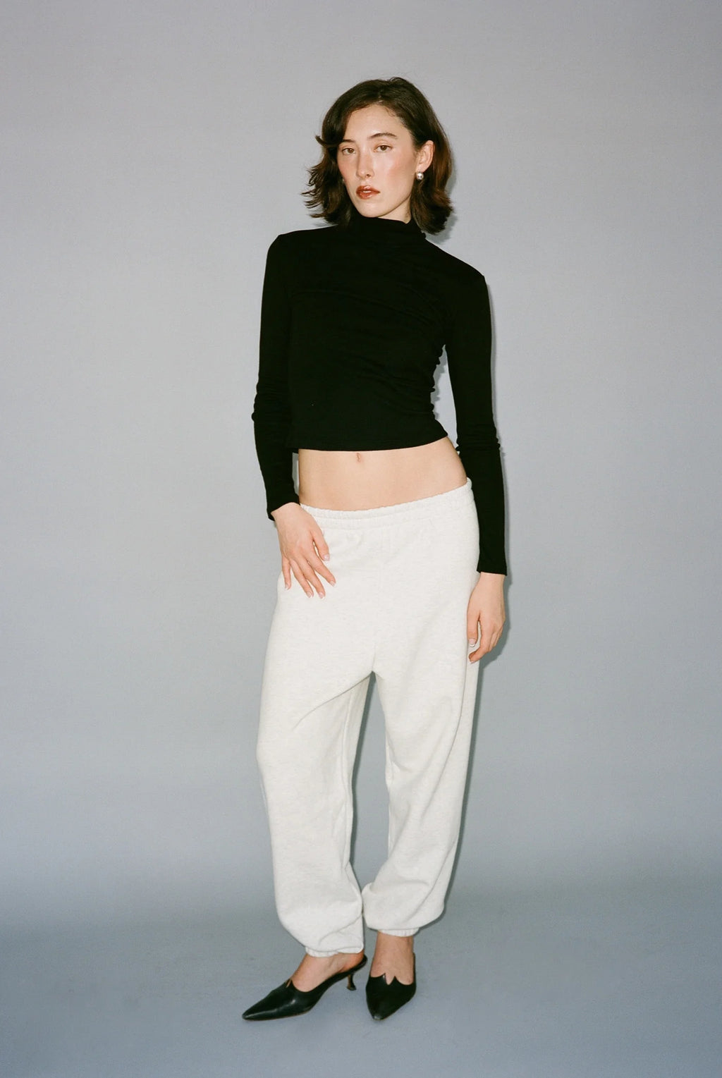 Woman wearing a black long-sleeve top and white pants against a gray background