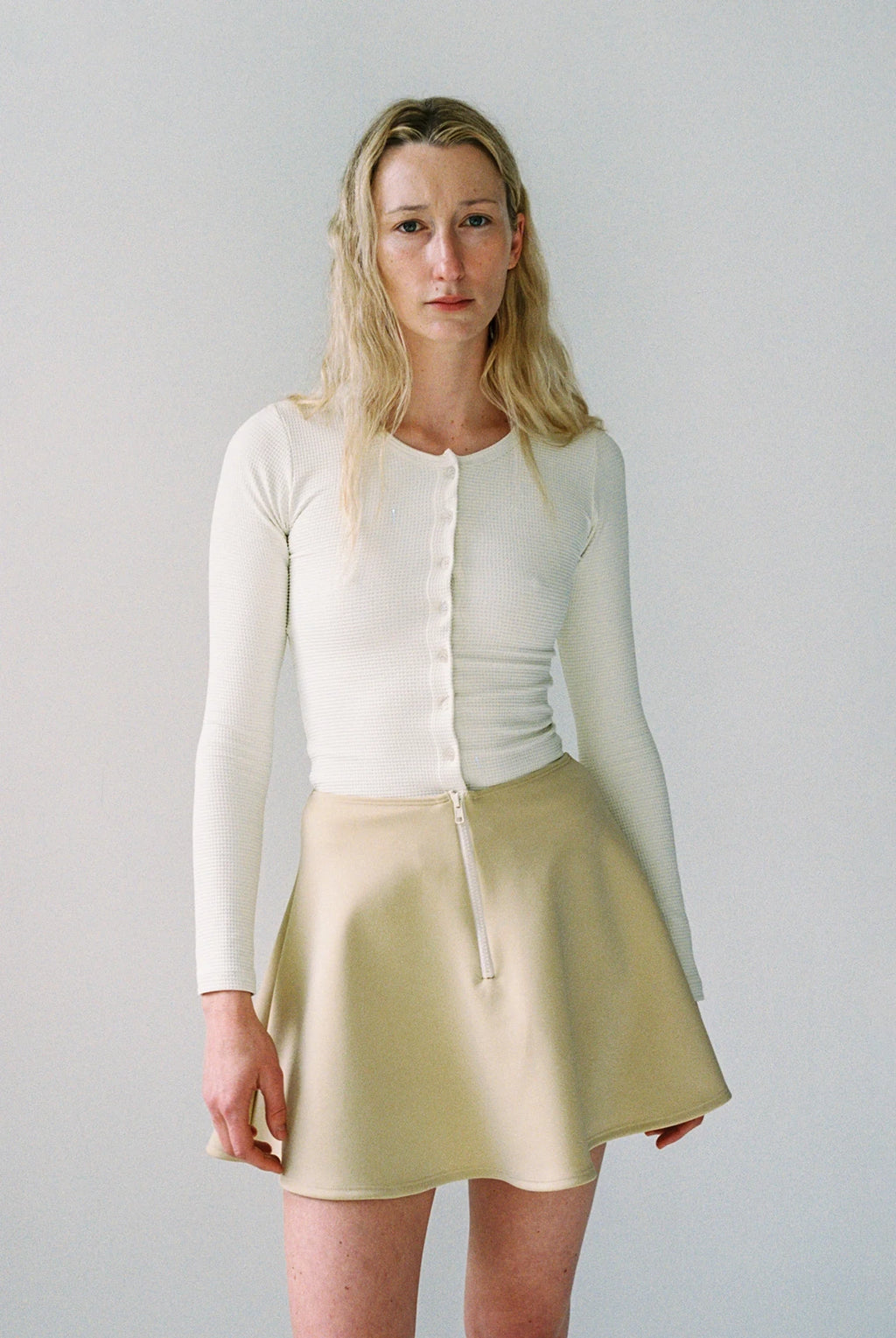 Woman wearing a white long-sleeve top and beige skirt against a plain background