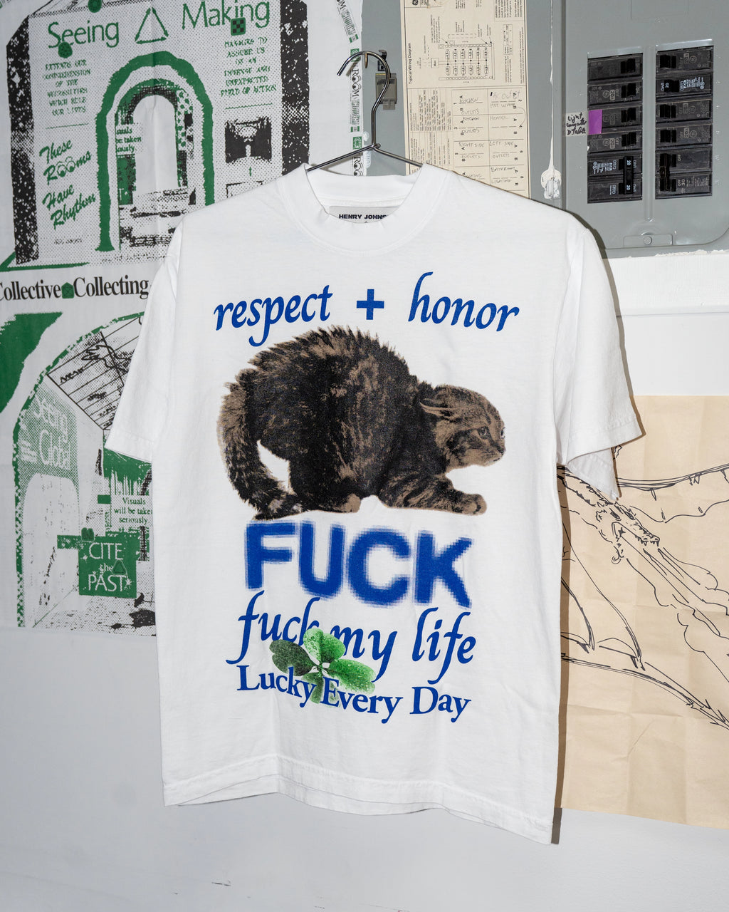 White t-shirt with graphic design and text on a hanger against a wall with posters.