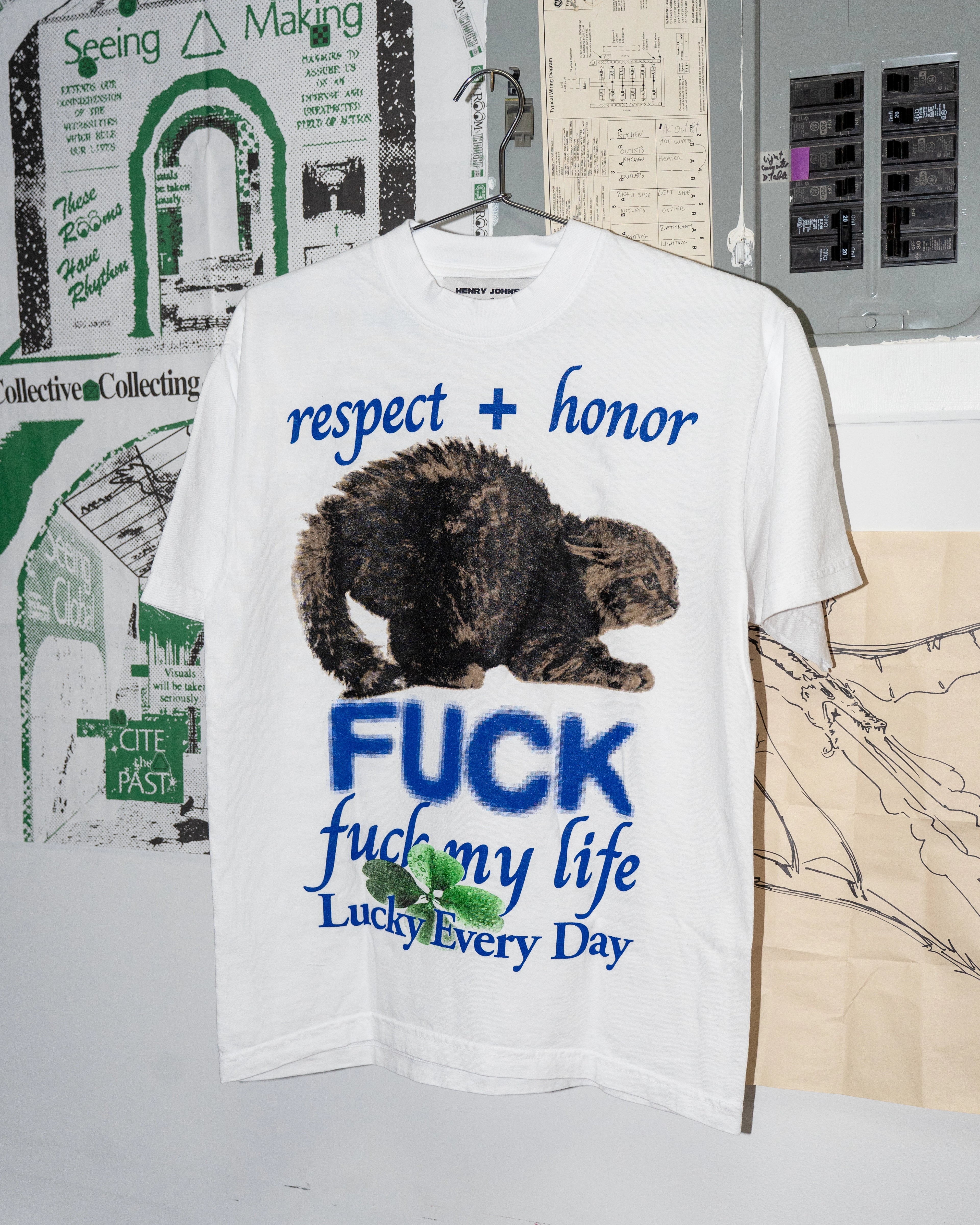 White t-shirt with graphic design and text on a hanger against a wall with posters.