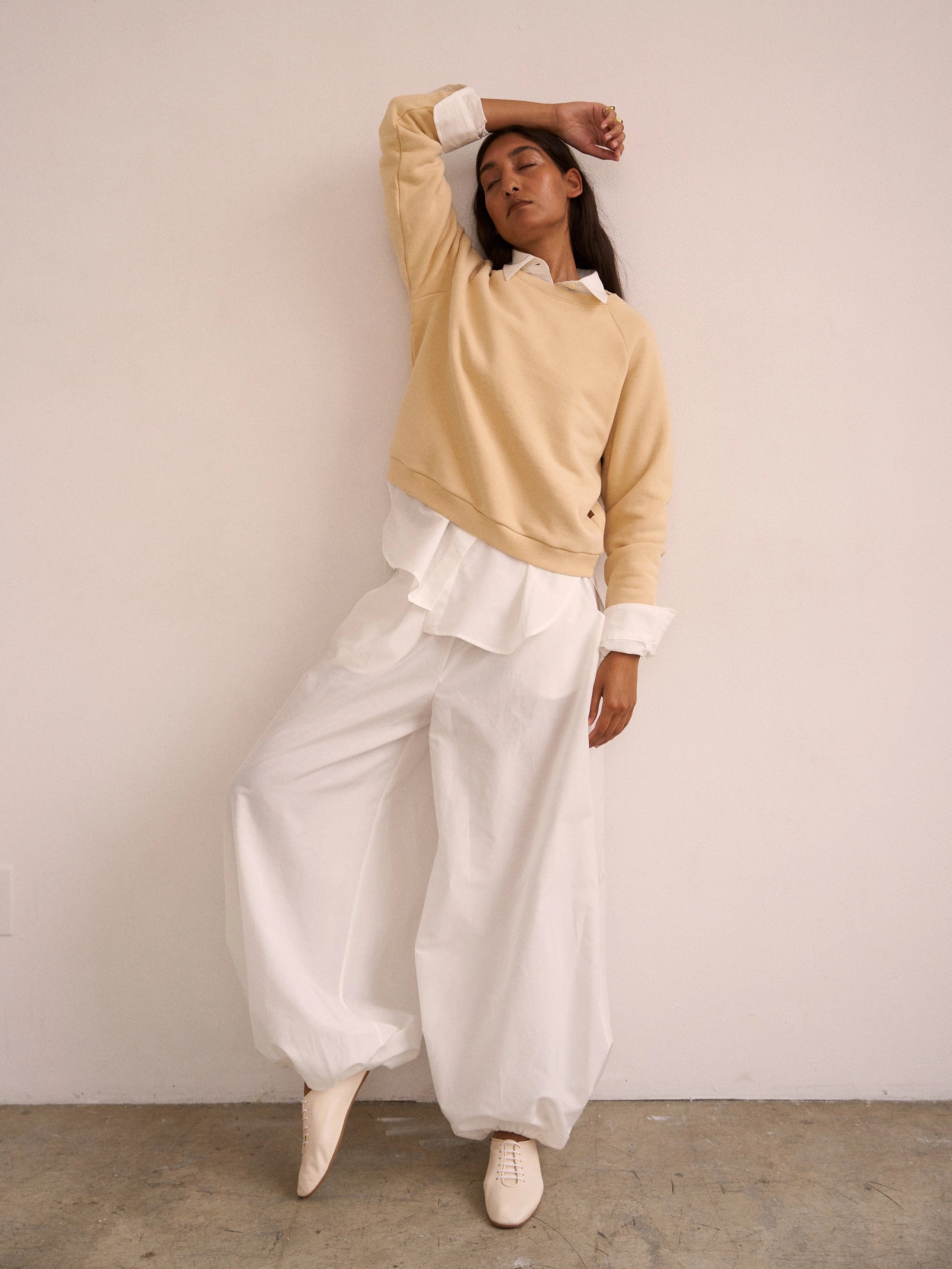 Woman wearing a beige sweater and white pants against a plain background