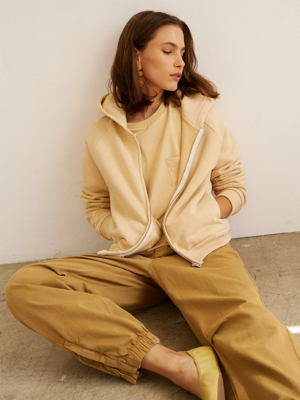 Woman wearing a beige tracksuit sitting on the floor against a plain wall.