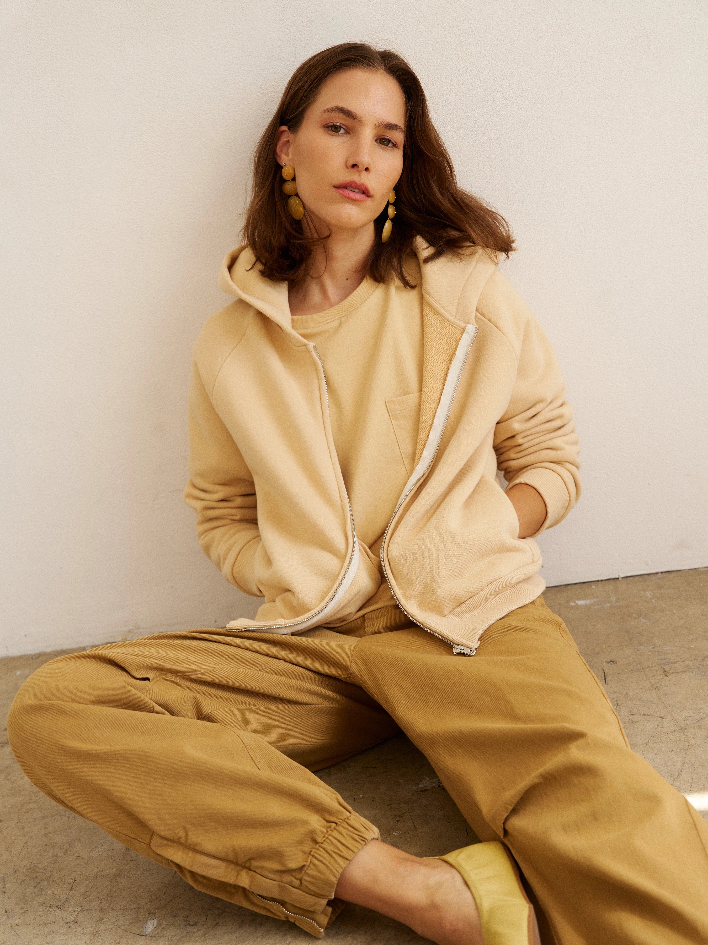 Woman wearing a beige tracksuit sitting on the floor against a white wall.