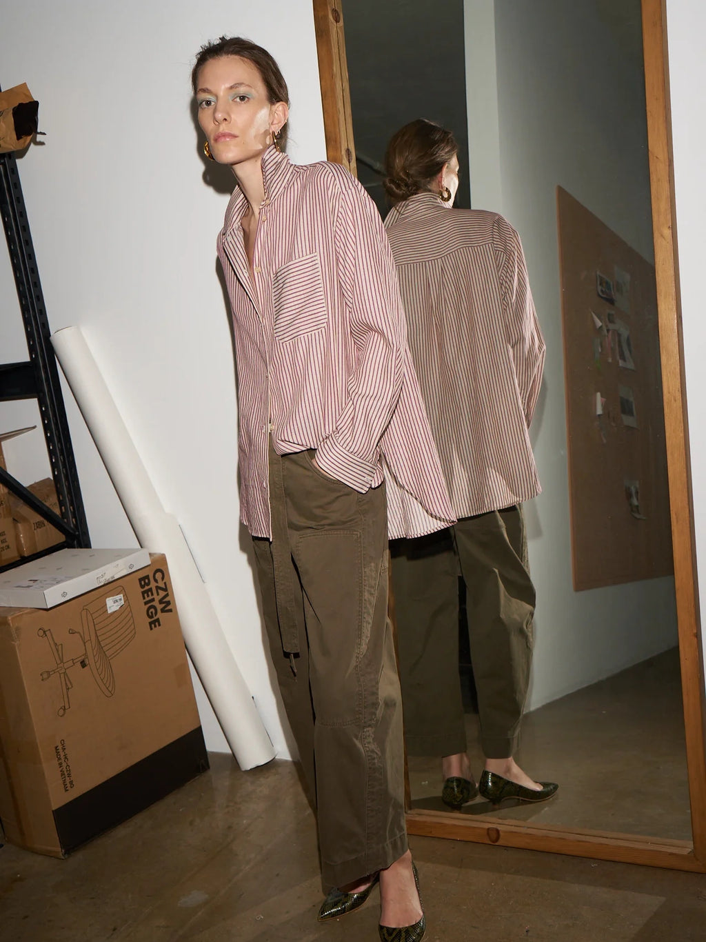 Woman wearing a pink striped shirt and green pants standing in front of a mirror.