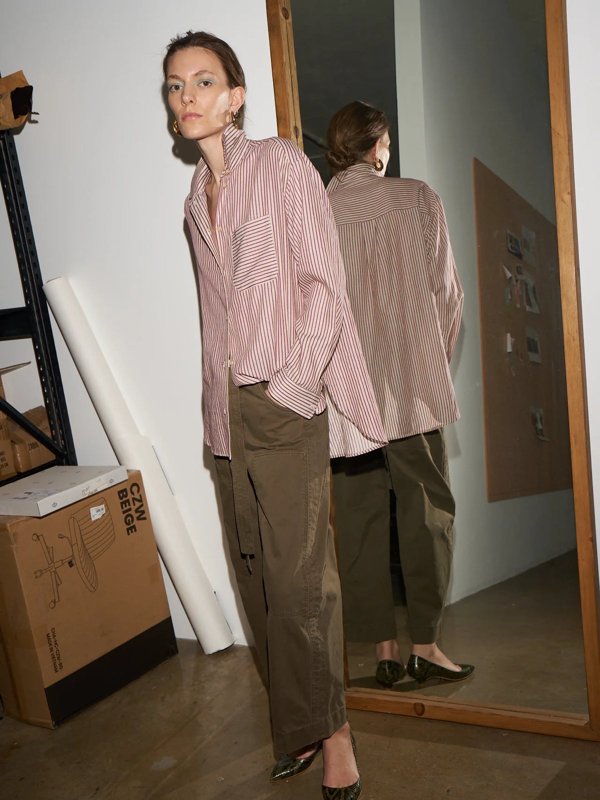Woman wearing a pink striped shirt and green pants standing in front of a mirror.