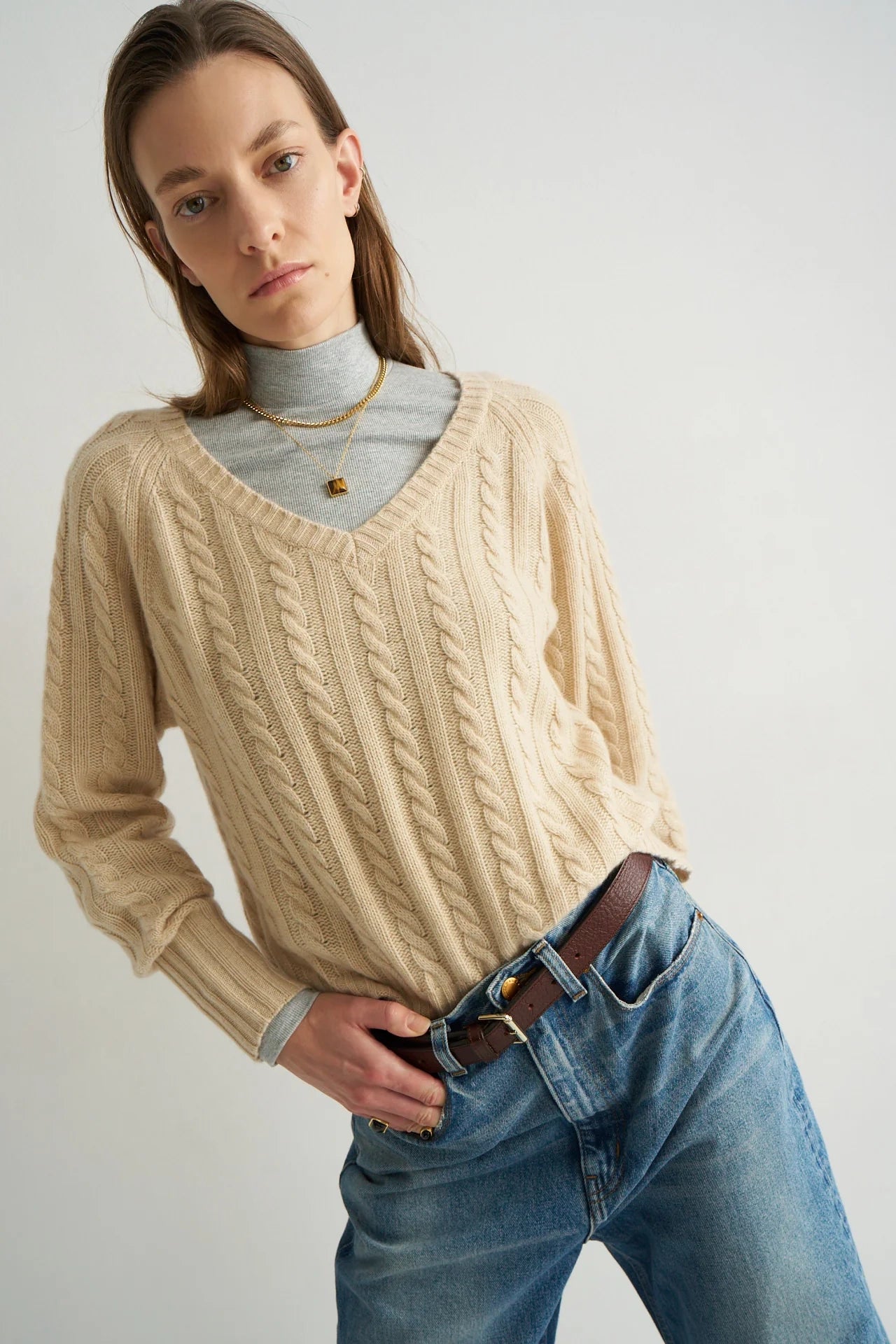 Person wearing a beige cable knit sweater over a turtleneck with blue jeans on a plain background