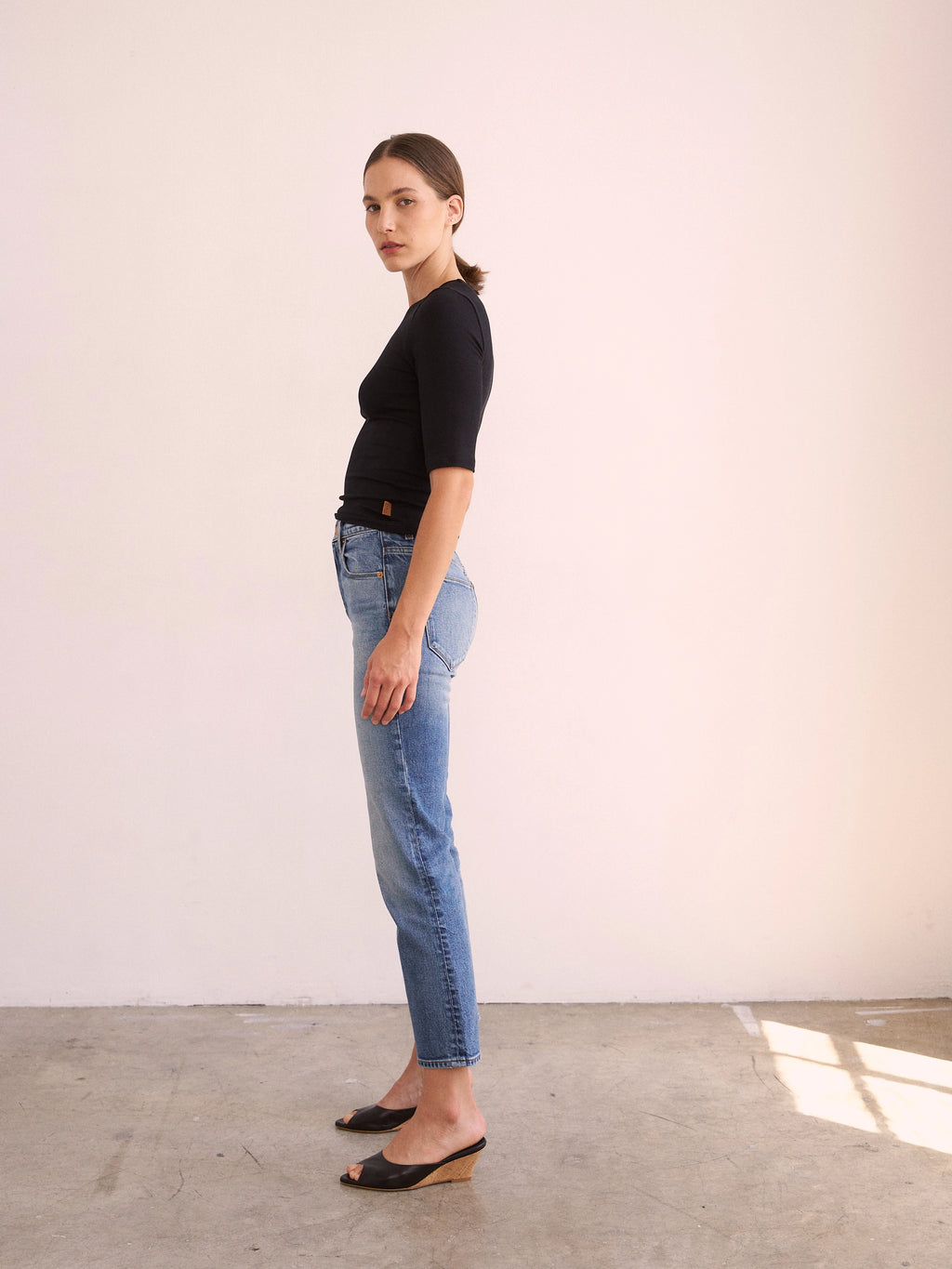 Woman wearing a black top and blue jeans standing against a white wall.