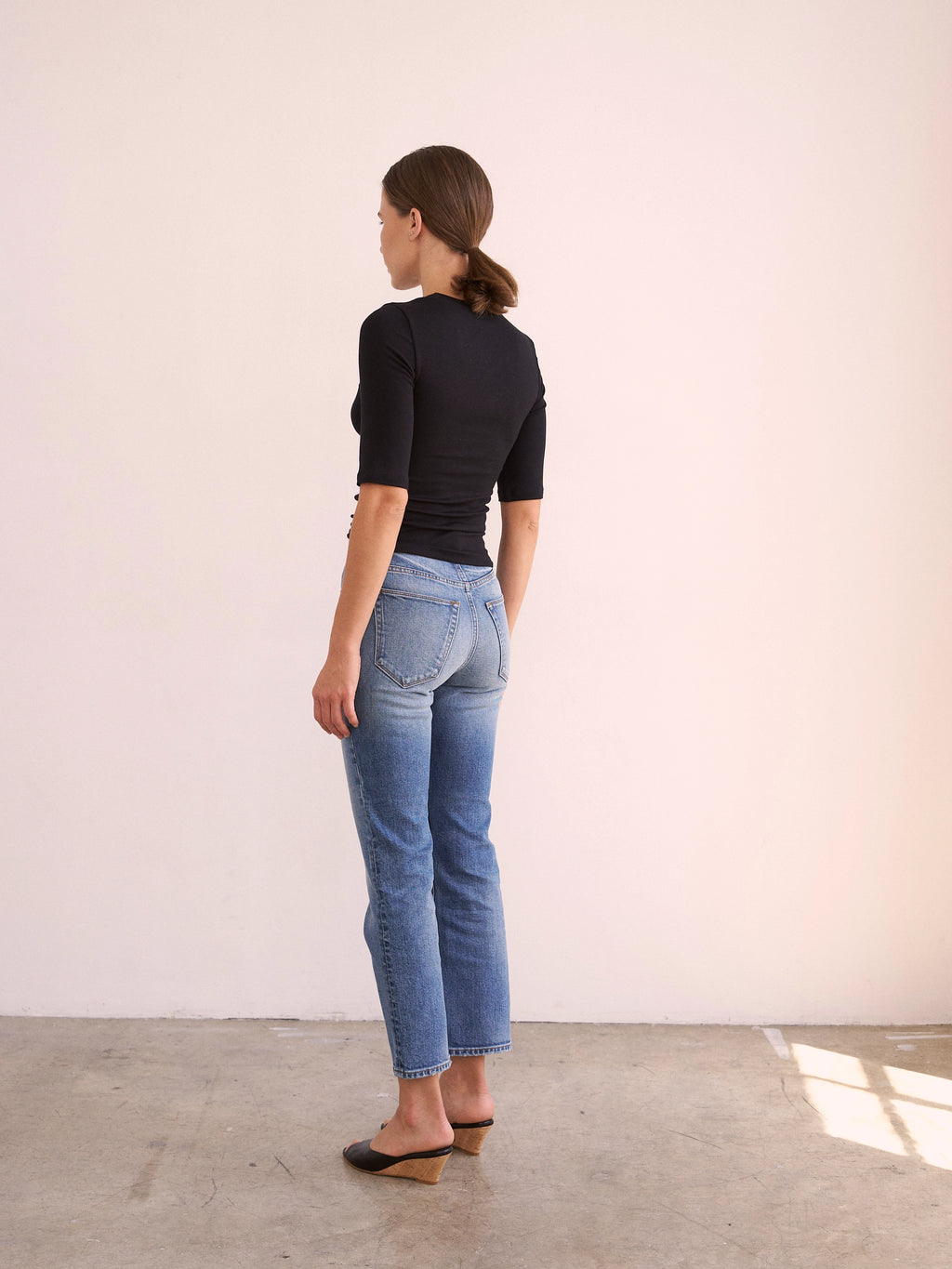 Woman wearing a black top and blue jeans standing against a white wall.