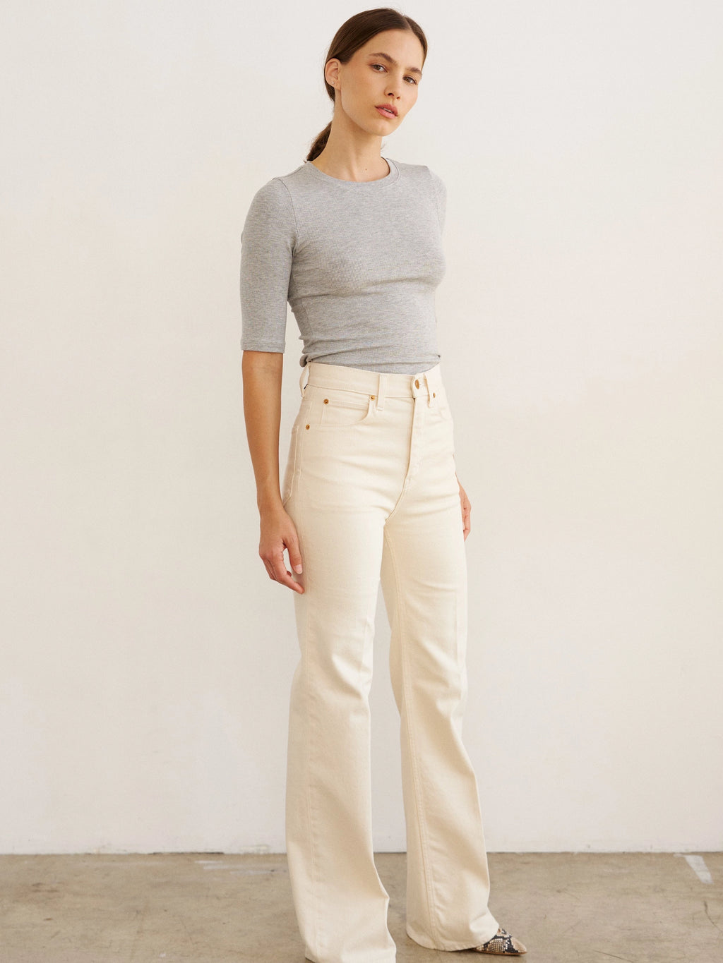 Woman wearing a gray sweater and white pants against a plain background