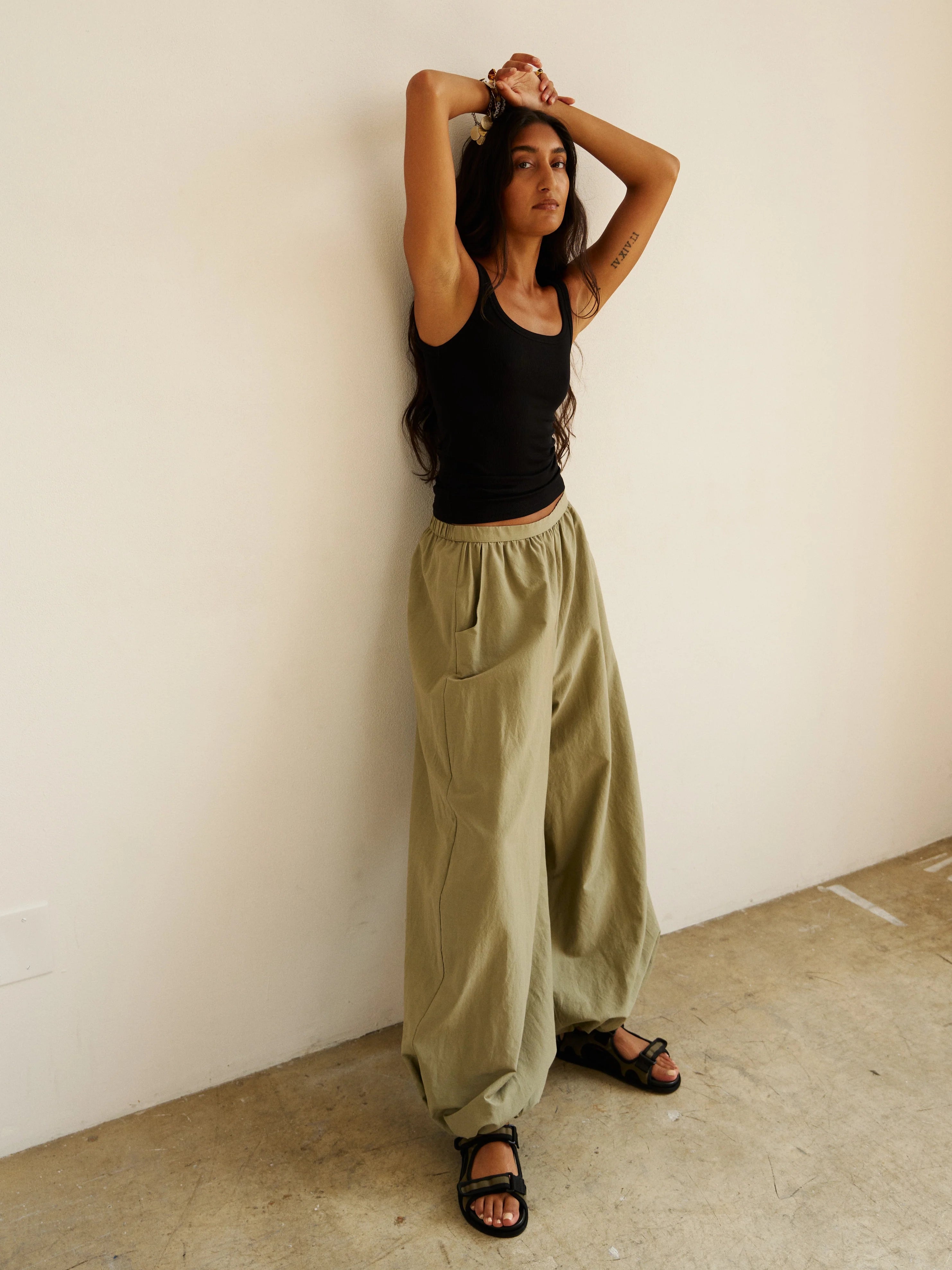 Woman wearing a black tank top and olive green pants against a white wall.