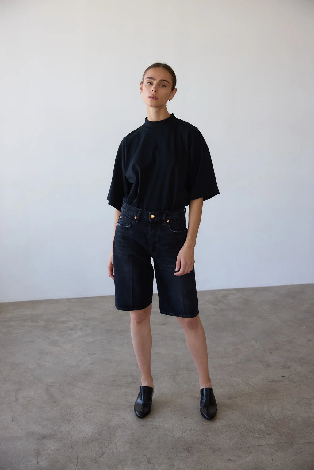 Person wearing a black t-shirt and dark denim shorts standing against a plain white wall.