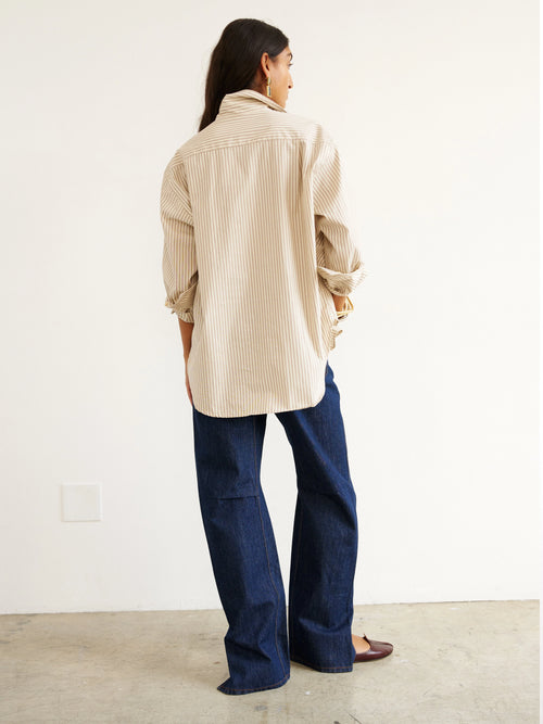 Person wearing a beige ribbed sweater and blue jeans standing against a white wall.