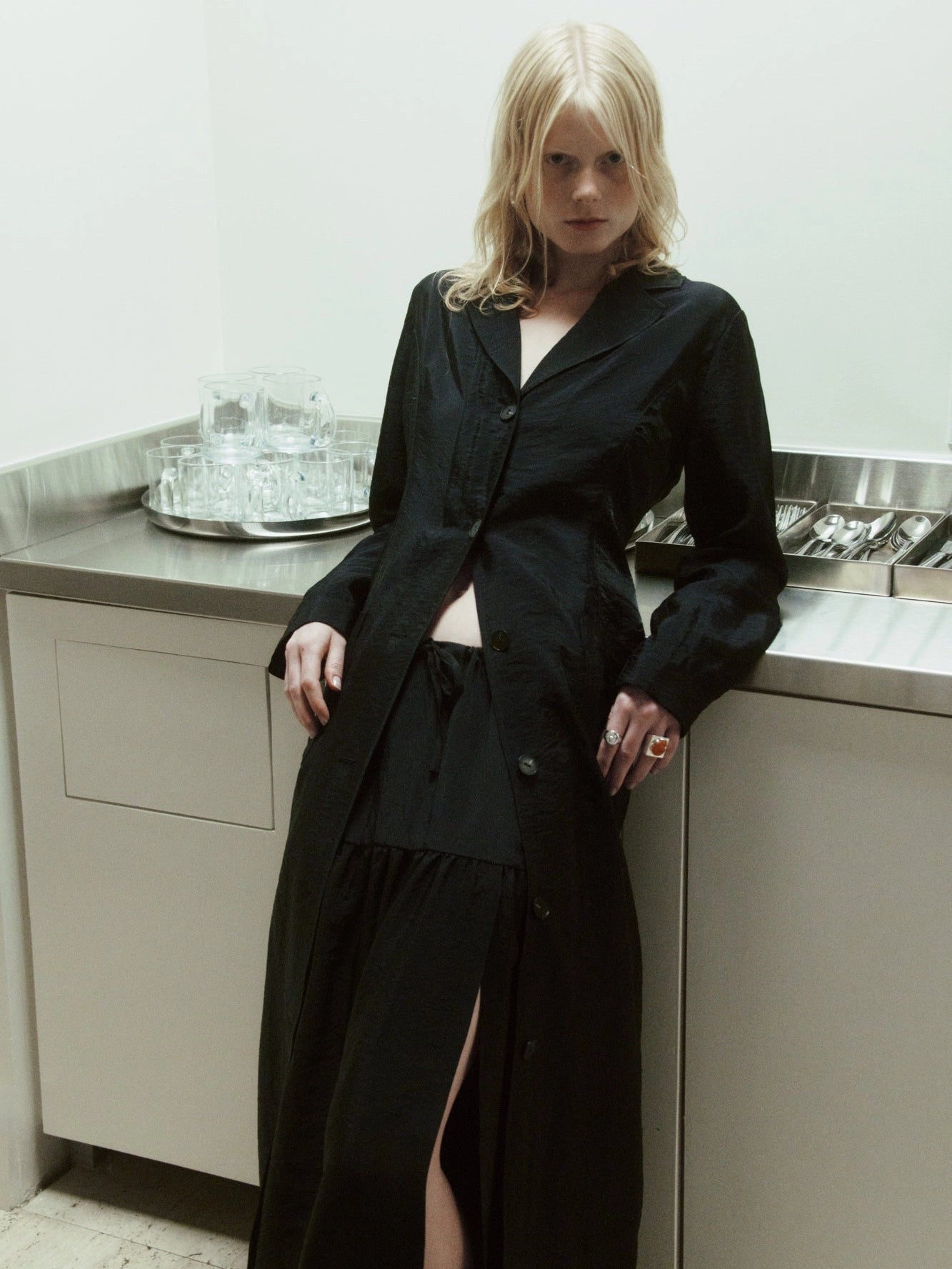 NOMIA duster coat in black with a straight, elongated silhouette, button front and back vent, worn over a matching skirt.