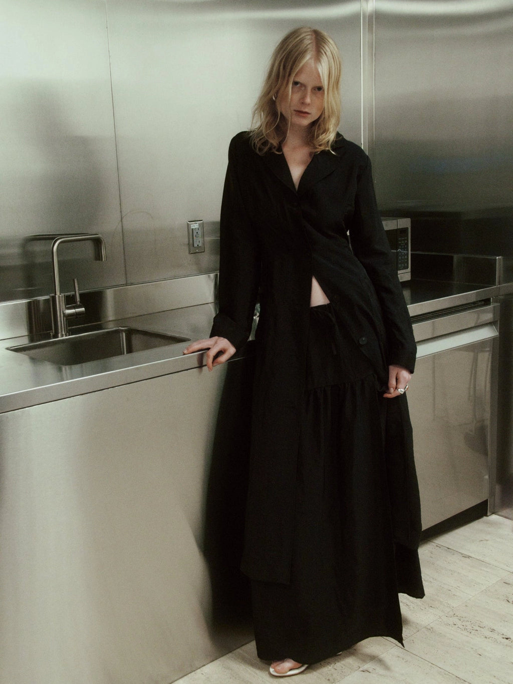 NOMIA duster coat in black with a straight, elongated silhouette, button front and back vent, worn over a matching skirt.