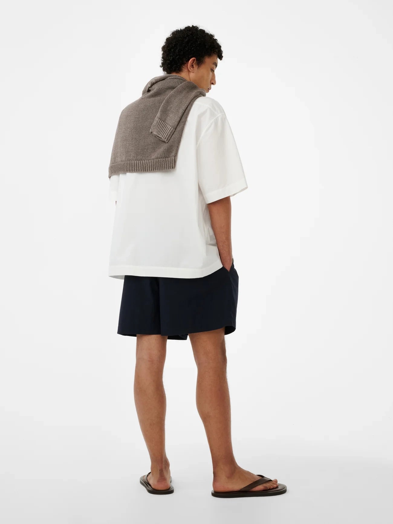 Person wearing a white shirt, black shorts, and a brown sweater over their shoulder on a white background