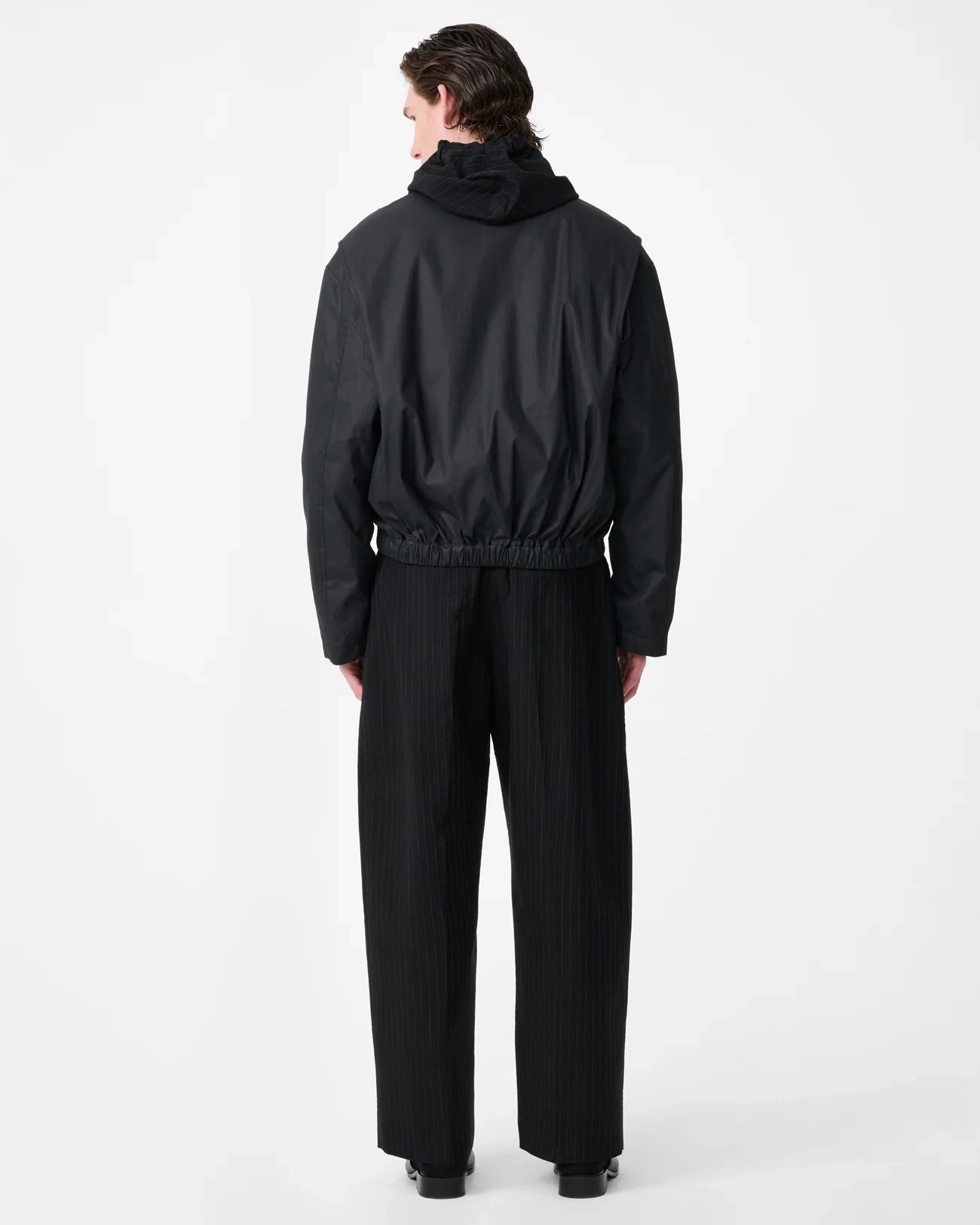 Model wearing Studio Nicholson Lyons black resinated cotton jacket with a classic collar, two-way zip front and elasticated hem.