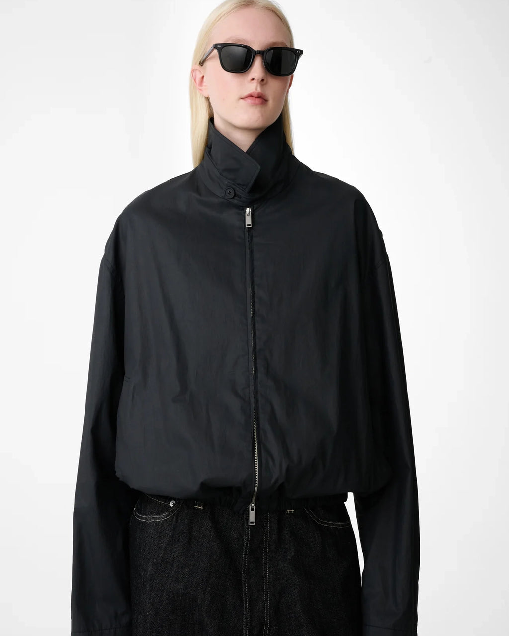 Model wearing Studio Nicholson Lyons black resinated cotton jacket with a classic collar, two-way zip front and elasticated hem.