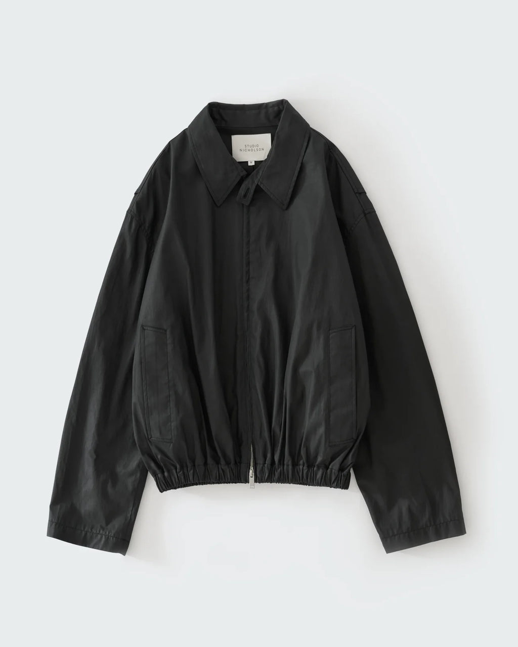 Studio Nicholson Lyons black resinated cotton jacket with a classic collar, two-way zip front and elasticated hem.