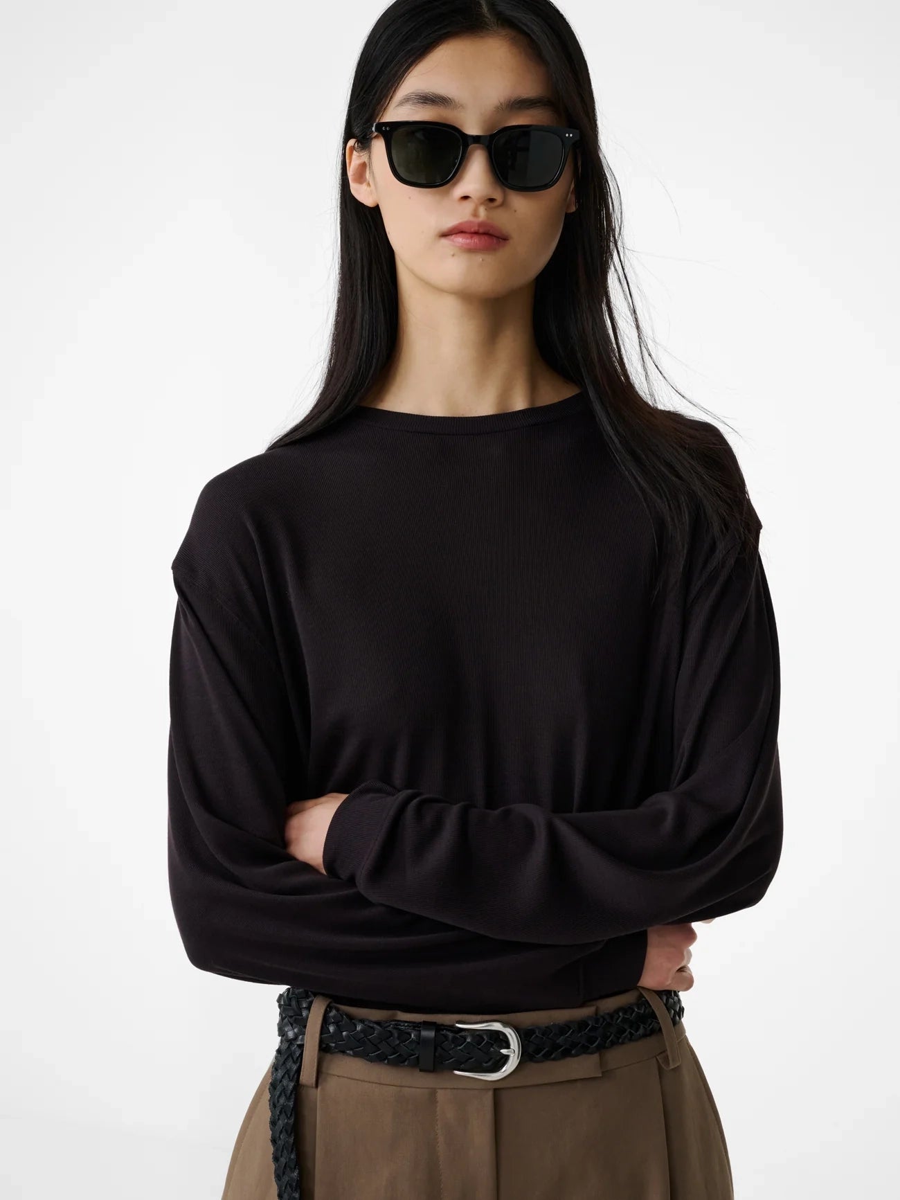 Woman wearing a black long-sleeve top and brown pants with a belt on a white background